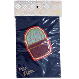 An embroidered cactus patch in a pot, packaged in a clear plastic bag with a branded header card. The patch features a green cactus with pink details and a dark brown pot, set against a dark navy blue backing card with gold foil text and a patterned top.