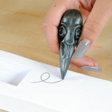 Hand holding a decorative graphite object with a bird-skull design, writing on a piece of paper.