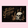 Black postcard with an ivory & gold illustration of a skull and candles, and the legend "Season's Greetings"