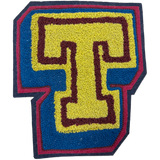 A hand-made chainstitch patch shaped like the letter T, featuring a yellow center with a dark burgundy outline, surrounded by a blue border and a dark navy outer edge.