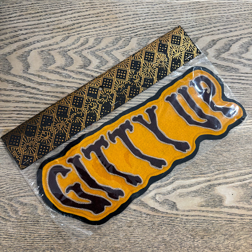 A yellow and black embroidered patch with the text 'CITY UP' in stylized lettering, shown next to its black and gold patterned backing paper on a gray wood-grain surface.