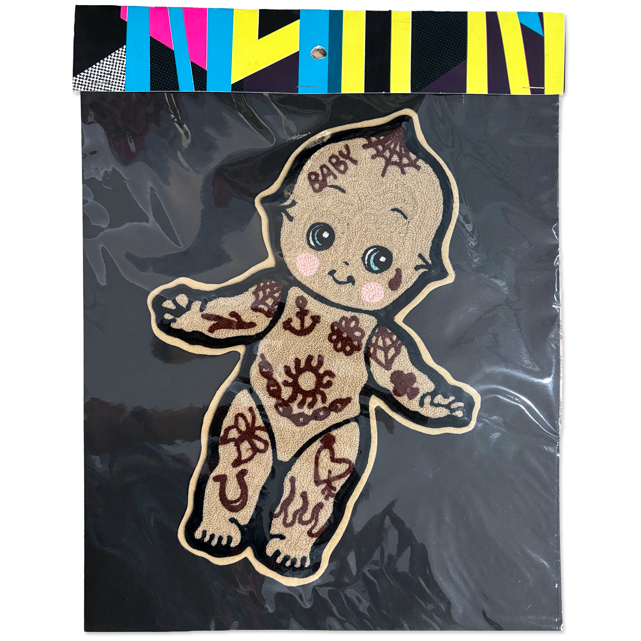 A decorative embroidered patch shaped like a baby figure, shown inside clear plastic packaging with a black backing and a colorful striped header card. The patch has a tan base with dark brown tattoo-style patterns and the word 'BABY' embroidered on the forehead.