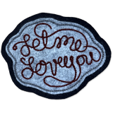 A felt patch with the phrase 'Let me love you' in dark brown cursive lettering, featuring a textured light blue center, a dark navy blue border, and a thin light blue inner border, shown on a plain white background.
