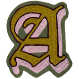 A hand-made chainstitch letter 'A' patch featuring a gold-yellow fill, light pink sections, a dark brown outline, and an olive green outer border, shown on a plain white background.