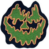 A handmade felt patch shaped like a jack-o'-lantern face, featuring textured green felt for the cheeks and forehead, black felt for the eyes and nose, and orange yarn trim outlining the design, set against a plain white background.