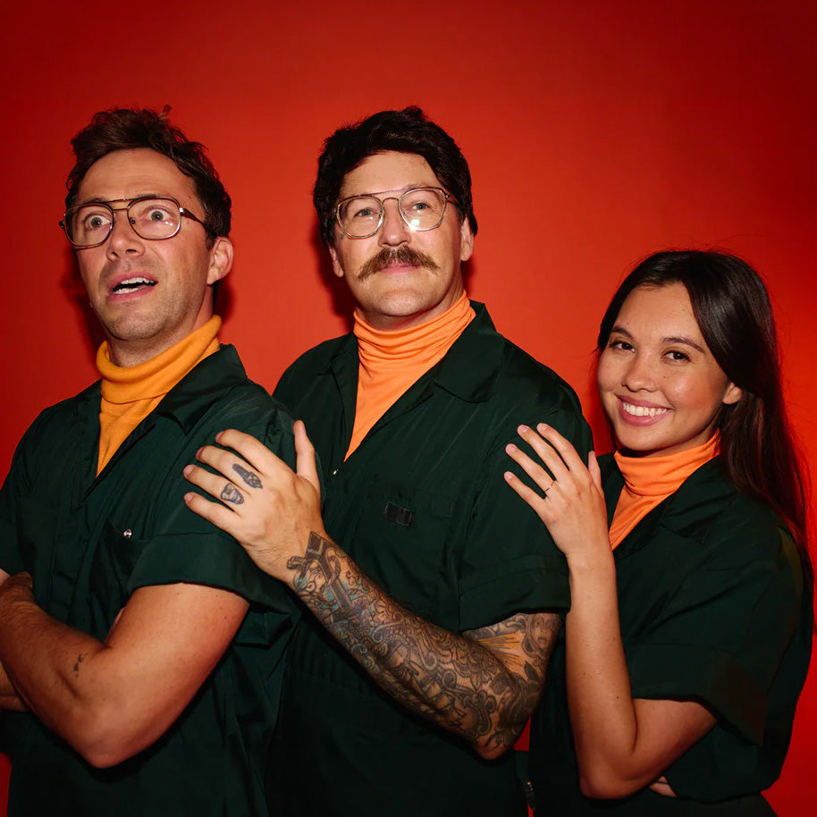 Three people posing together in matching dark green scrubs over bright orange turtlenecks, standing against a solid red-orange background.